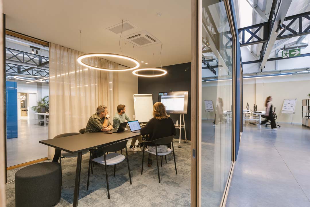 Drie collega's in overleg aan een vergadertafel in een modern kantoor met glazen wanden en ronde hanglampen.